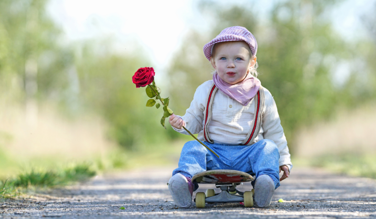 Ein kleiner, schick angezogener Junge mit einer Rose in der Hand sitzt auf einem Skateboard.