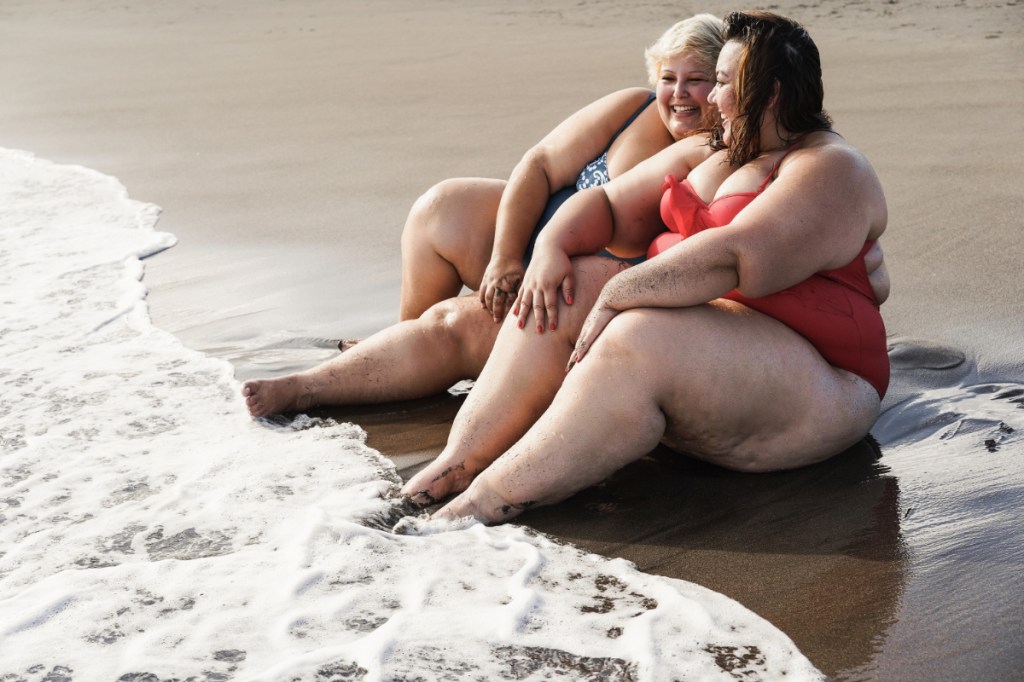 Zwei gut gelaunte, vollschlanke Frauen im Bikini sitzen am Strand am Wasser.