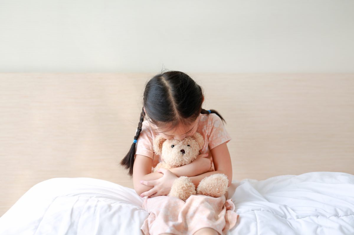 Ein kleines Mädchen sitzt auf ihrem Bett und umarmt ihr Stofftier.