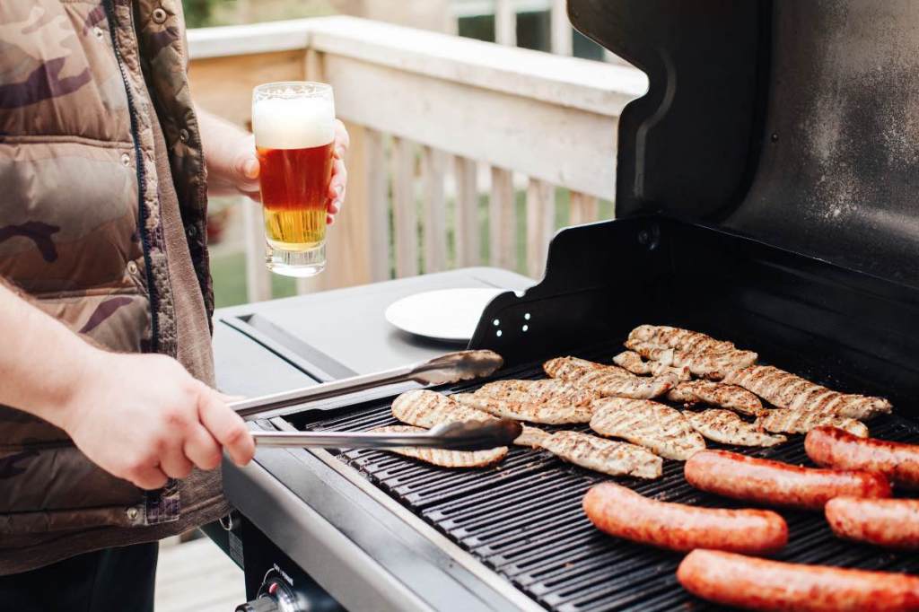 Ein Bild von einem Mann der mit einem Bier in der Hand und einer Grillzange Fleisch grillt.