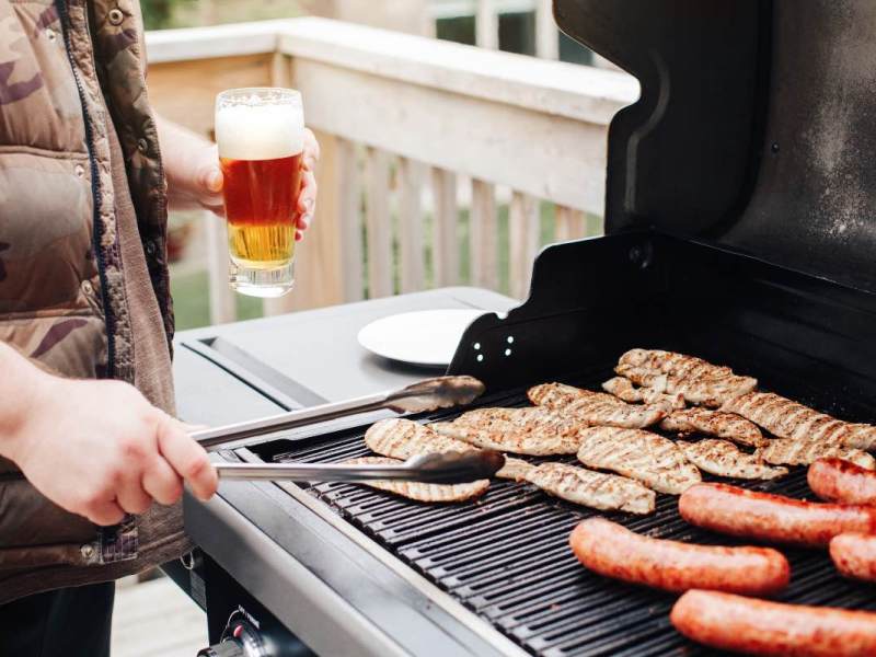 Ein Bild von einem Mann der mit einem Bier in der Hand und einer Grillzange Fleisch grillt.