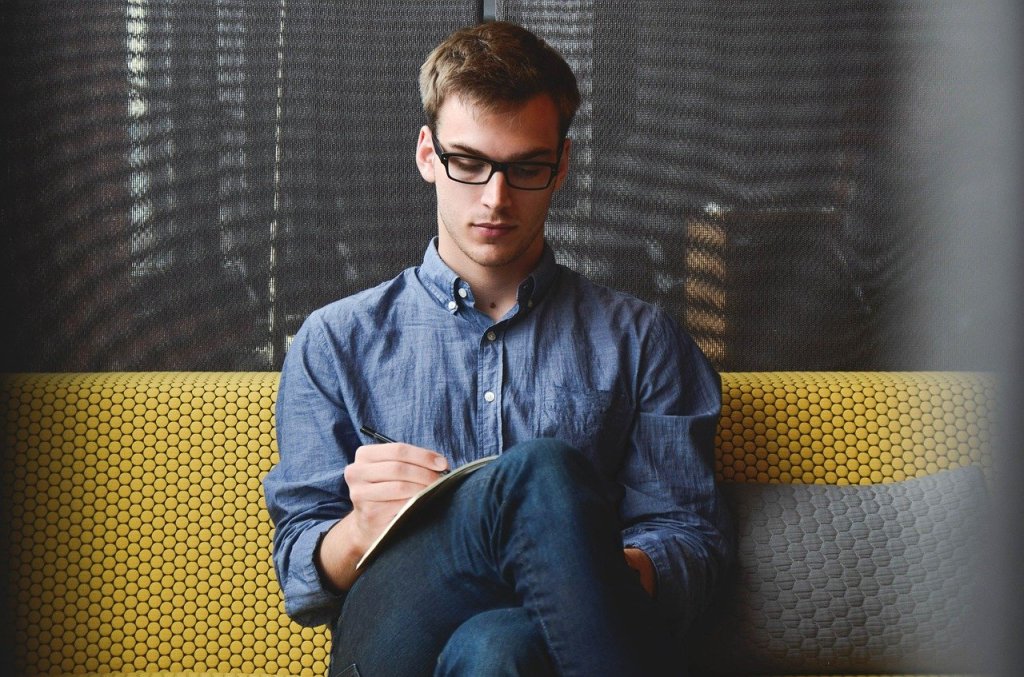 Ein junger Mann mit Brille sitzt auf dem Boden und schreibt etwas in ein Heft.