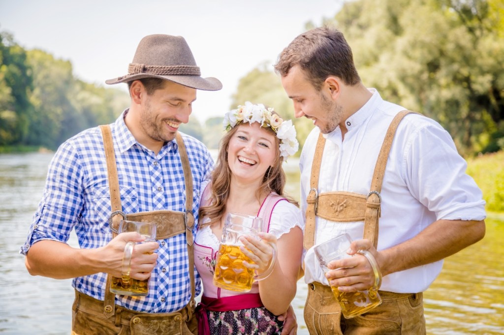 Drei Freunde in bayrischer Tracht stehen an einem Fluss und trinken Bier.