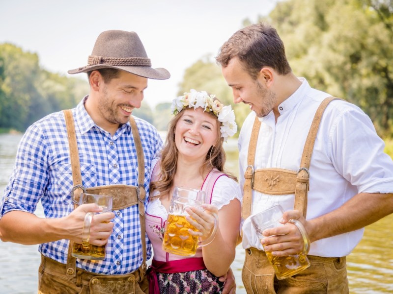 Drei Freunde in bayrischer Tracht stehen an einem Fluss und trinken Bier.