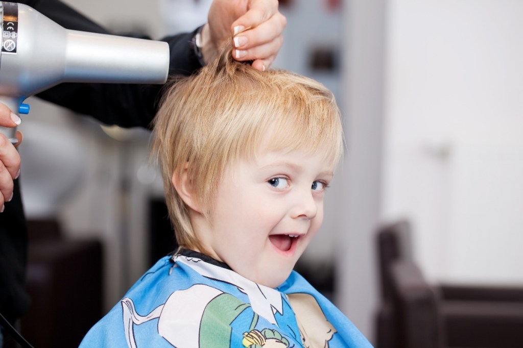 Ein blonder Junge bekommt beim Friseur die Haare geföhnt und lacht.