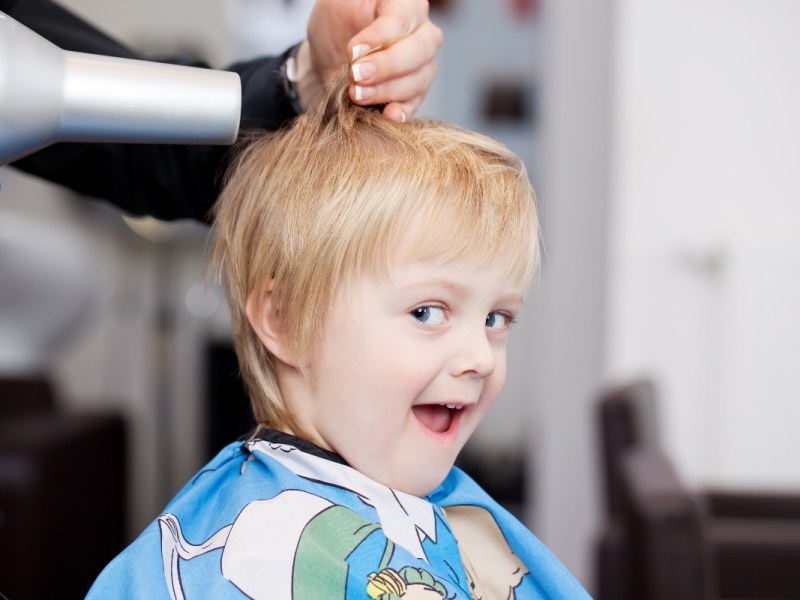 Ein blonder Junge bekommt beim Friseur die Haare geföhnt und lacht.