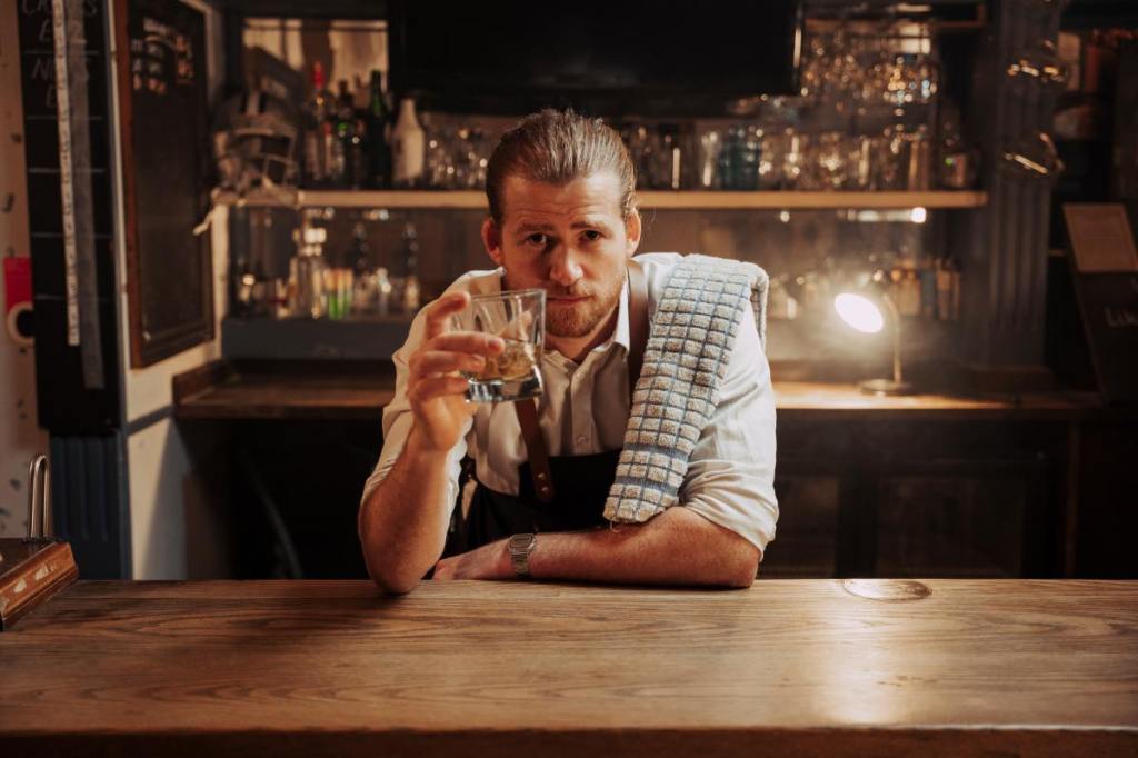 Ein junger Wirt trinkt hinter seinem Tresen einen Whiskey nach Feierabend.