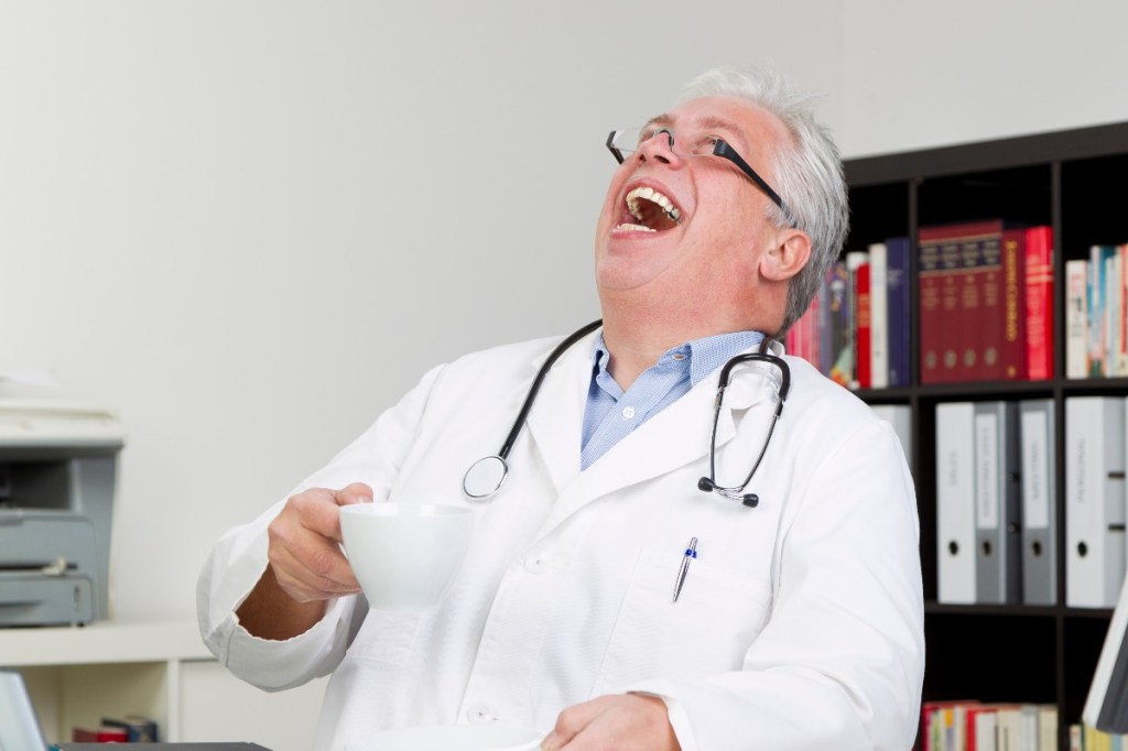 Ein älterer Arzt im weißen Kittel mit einer Kaffeetasse in der Hand lacht herzhaft.