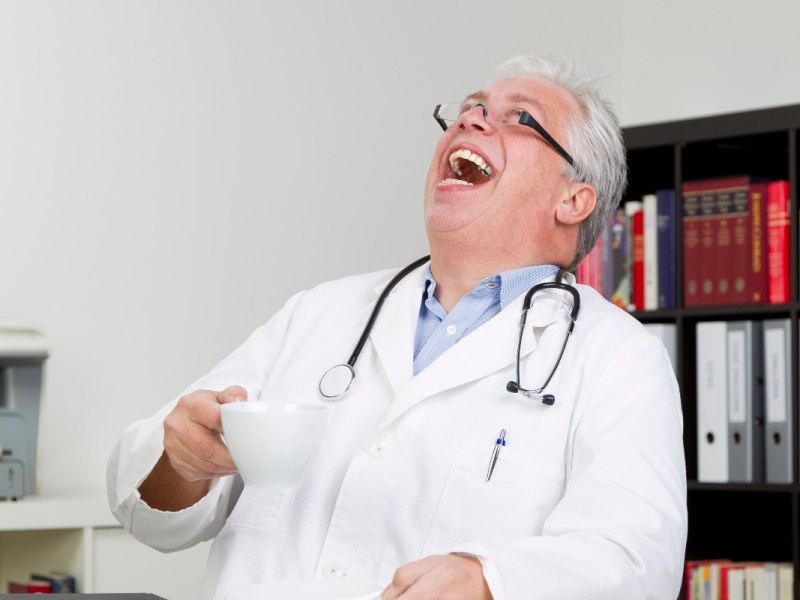 Ein älterer Arzt im weißen Kittel mit einer Kaffeetasse in der Hand lacht herzhaft.