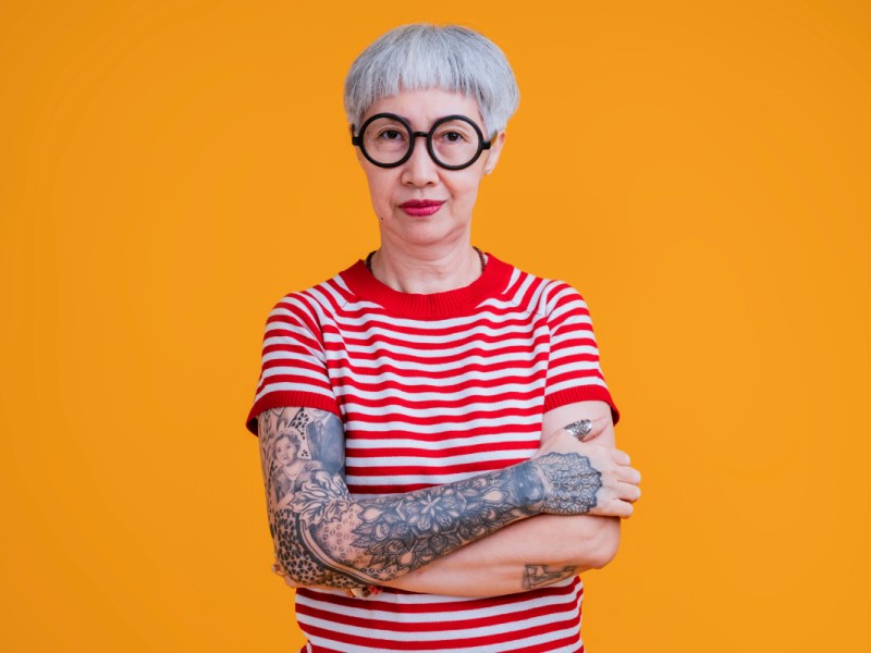Eine ältere Frau mit Brille, T-Shirt und tätowierten Armen steht vor einem orangenem Hintergrund.