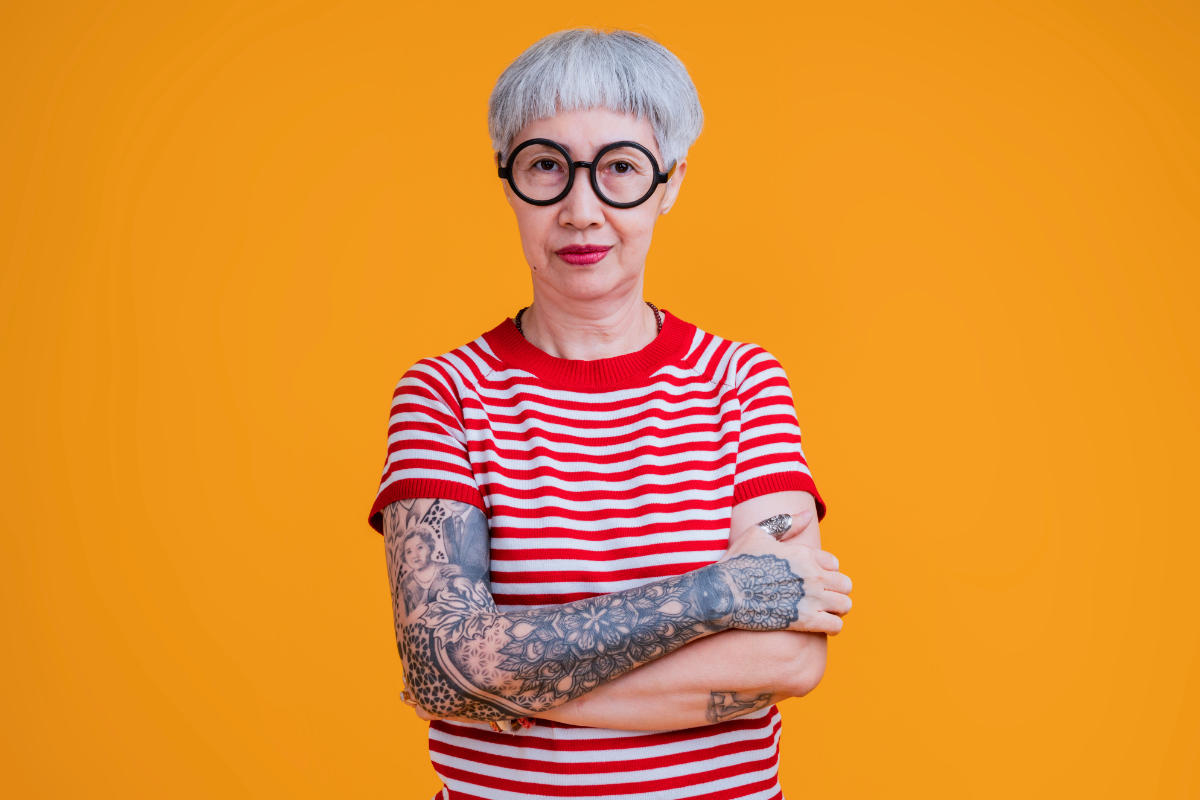 Eine ältere Frau mit Brille, T-Shirt und tätowierten Armen steht vor einem orangenem Hintergrund.