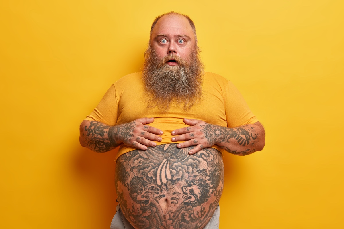 Ein bärtiger Mann zeigt seinen tätowierten Bauch, vor gelbem Hintergrund.