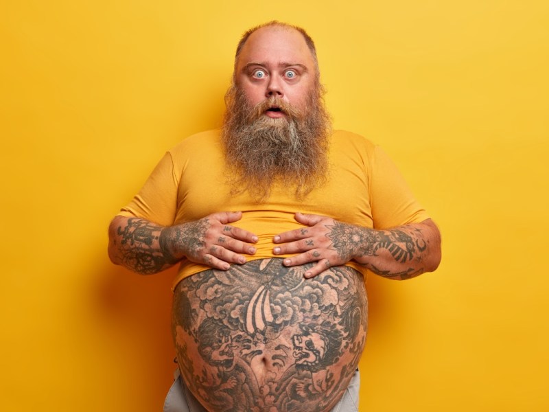 Ein bärtiger Mann zeigt seinen tätowierten Bauch, vor gelbem Hintergrund.