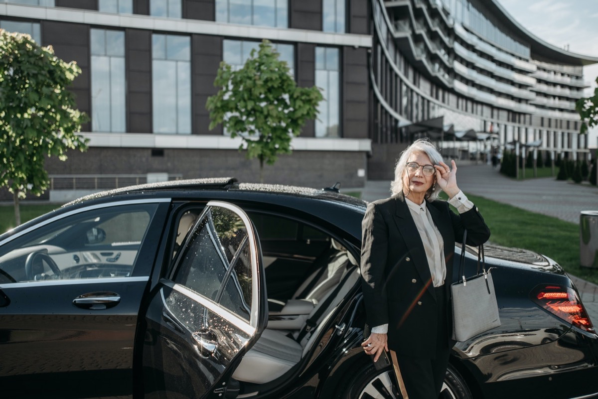 Eine alte Frau mit grauen Haaren steht vor der offenen Tür ihres Autos.