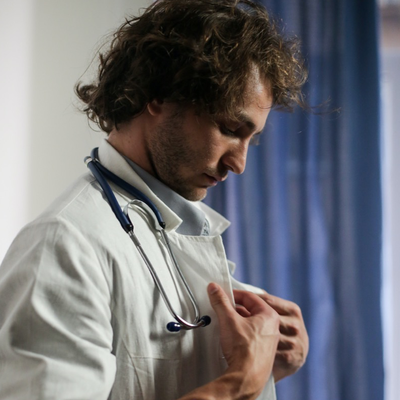 Ein junger Arzt mit Stethoskop knöpft sich den weißen Kittel zu