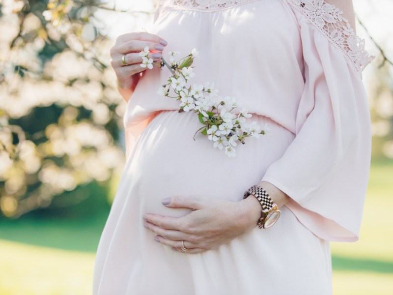 Eine schwangere Frau hält sich den Bauch auf einer Wiese im Frühling.