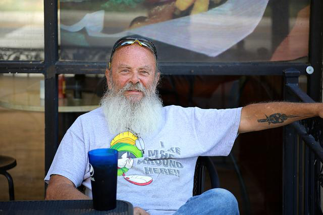 Ein alter Mann mit Bart und Taetowierung und Sonnenbrille trinkt eine Cola