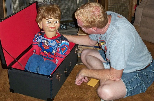 Ein Bauchredner holt eine Marionette mit Superman-Kostüm aus einer Kiste