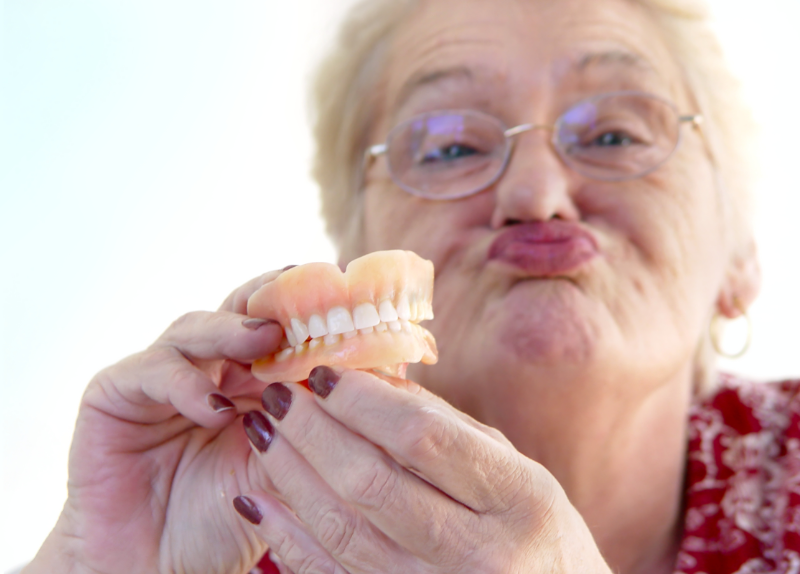 Eine alte Frau mit Brille hält ihr Gebiss in den Händen und macht einen Schmollmund