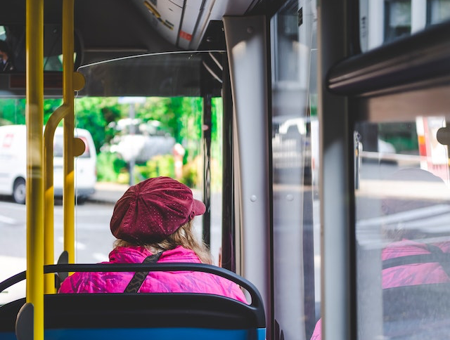 Eine alte Frau mit roter Mütze und pinker Jacke sitzt im Bus.