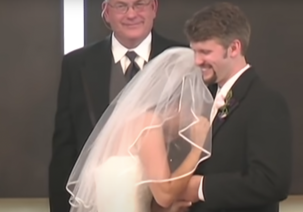 Eine Braut liegt ihrem lachenden Ehemann in den Armen und der Pastor lächelt