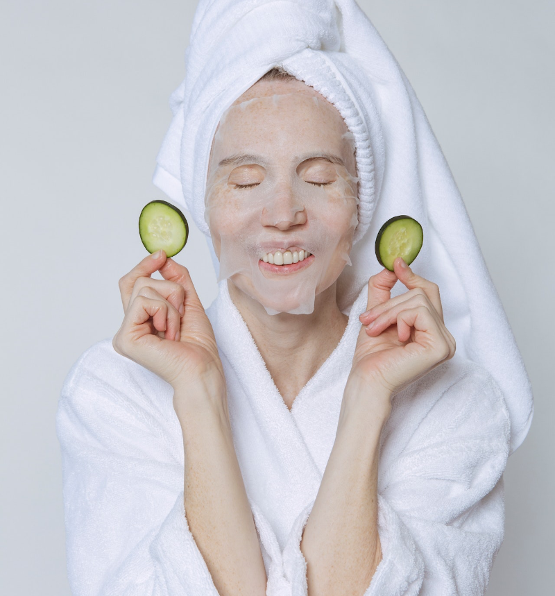 Eine Frau, mit Handtuch um den Kopf gewickelt, trägt eine Schönheitsmaske mit Gurkenscheiben auf