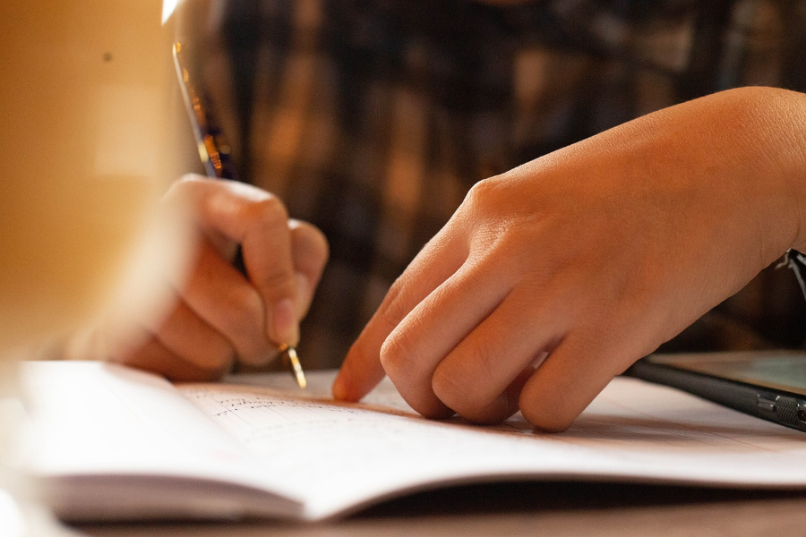 Man sieht die Hände eines Schülers, der seine Hausaufgaben macht. Er schreibt etwas in sein Heft.